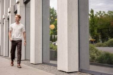 Kızıl saçlı, yirmili yaşlarda bir elinde dizüstü bilgisayar taşıyan bir adam binanın etrafında hareket ediyor.