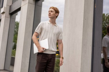 Genç kızıl adam kapalı dizüstü bilgisayarla şehir binasının yakınında yürüyor ve başka tarafa bakıyor.