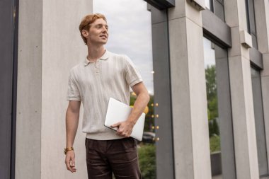 Kızıl saçlı genç erkek cam ofis binasının yanında dizüstü bilgisayar tutuyor.