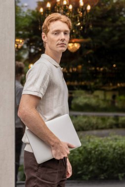 Kızıl saçlı adam ofis binasının önünden geçiyor ve elinde dizüstü bilgisayarla yeşil çiçek tarhlarından geçiyor.