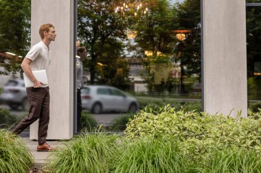 Kızıl saçlı genç erkek cam ofis binasının yanında dizüstü bilgisayar tutuyor. Etrafı bitkilerle çevrili.