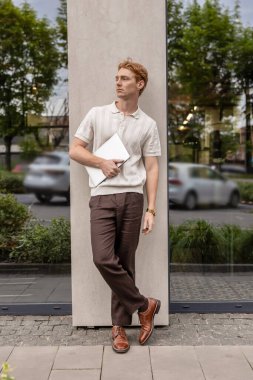 Kızıl saçlı adam elinde dizüstü bilgisayarla sokak duvarının yanında duruyor.