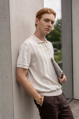 20 'li yaşlarda genç bir kızıl, elinde kapalı bir dizüstü bilgisayarla dışarıda ofis binalarının yanında duruyor.