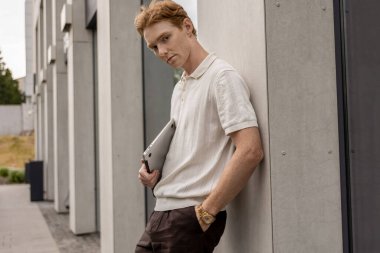 Modern ofis binasının yanında duran kızıl saçlı erkek elinde dizüstü bilgisayarla kameraya bakıyor.