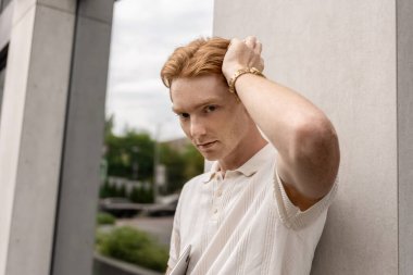 Genç kızıl erkek, duvarın yanında duruyor. Elinde dizüstü bilgisayar var.