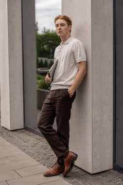 Genç kızıl erkek, duvarın yanında duruyor. Elinde dizüstü bilgisayar var.