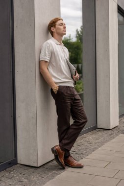 Kızıl saçlı adam dışarıda duvar inşa ederken bilgisayarını kapatmış poz veriyor.