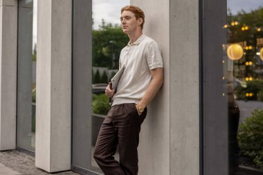 Kızıl saçlı, genç bir yetişkin, bir elinde dizüstü bilgisayarla duvara yaslanıyor ve ileri bakıyor.