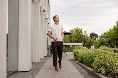 Kızıl saçlı genç bir erkek elinde bilgisayarla dışarıda yürüyor. Etrafı bitkilerle çevrili.