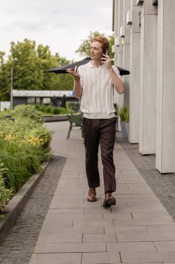 Genç kızıl adam ofis binasından caddeye doğru yürüyor ve akıllı telefondan konuşuyor.