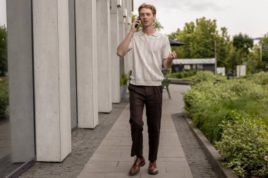 Kızıl adam ofis binasının önünden geçiyor ve dışarıda dolaşırken cep telefonuyla konuşuyor.