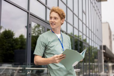 Cam klinik binasının yanında dikilen laboratuvar önlüklü kızıl doktor elinde klinik geçmişi olan bir dosya tutuyor.