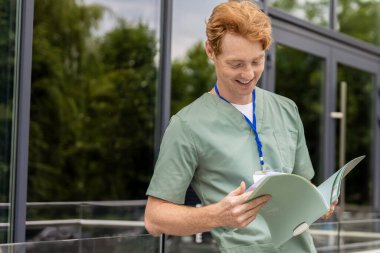 Laboratuvar önlüklü, kızıl saçlı sağlık çalışanı cam binanın yanında hasta geçmişi olan belgeleri tutuyor.