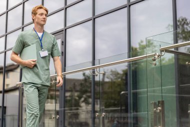 Laboratuvar önlüklü kızıl doktor cam klinik binasının önünden geçerken elinde klinik geçmişi olan bir dosya tutuyor.