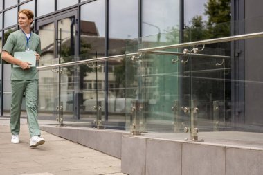 Laboratuvar önlüklü kızıl saçlı sağlık çalışanı hasta geçmişi olan cam binanın önünden geçiyor.