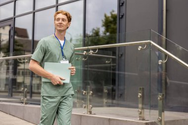 Laboratuvar önlüklü kızıl doktor cam klinik binasının önünden geçerken elinde klinik geçmişi olan bir dosya tutuyor.