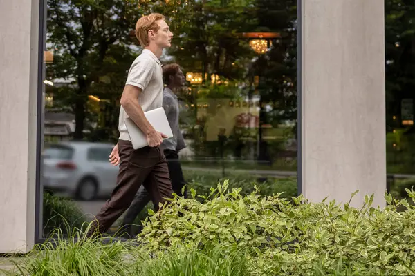 Kızıl saçlı adam ofis binasının önünden geçiyor ve elinde dizüstü bilgisayarla yeşil çiçek tarhlarından geçiyor.