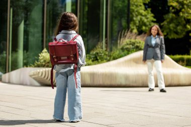 Kırmızı sırt çantalı genç bir kız şık bankın yanında gülümseyen bir kadına doğru yürüyor.