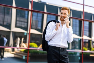 Beyaz tişörtlü kızıl saçlı adam omzunun üzerinde ceketini tutuyor ve modern şehir ortamında akıllı telefondan konuşuyor.