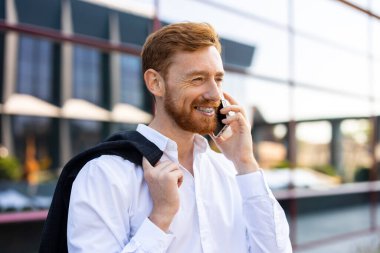 Beyaz tişörtlü kızıl saçlı adam dışarıda duruyor ve modern şehir ortamında akıllı telefondan konuşuyor.