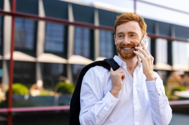 Kızıl sakallı adam iş bölgesinde cam binanın yanında cep telefonuyla konuşuyor.