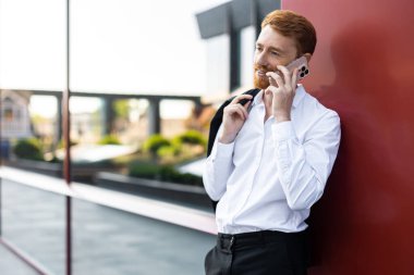 Sakallı, beyaz gömlekli, ceketli, ofis binasının önünde konuşan kızıl saçlı adam.