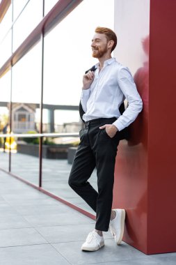 Beyaz gömlekli, siyah pantolonlu, kırmızı saçlı adam modern şehir binasının yanında ceketini tutuyor.