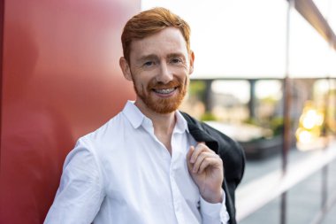 Kızıl sakallı, beyaz gömlekli ve resmi pantolonlu bir adam kırmızı bina duvarına karşı poz veriyor.