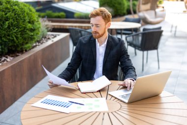 Bilgisayar üzerinde çalışırken basılı belgeleri yazıp kontrol eden kızıl saçlı bir profesyonel.