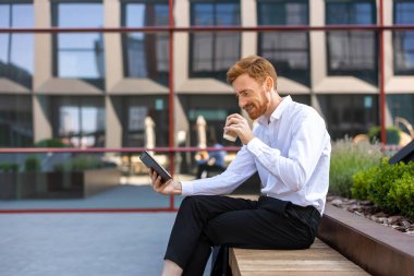 Sakallı gülen işadamı bankta oturup kahve içerken tablet kullanıyor.
