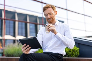 Beyaz gömlekli kızıl işadamı açık hava bankında kahve içerken tabletle web sayfalarını karıştırıyor.