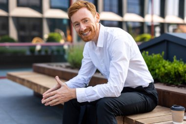 Beyaz gömlekli kızıl sakallı adam güneşli şehir sokağında bankta oturmuş kendinden emin bir ifadeyle ileriye bakıyor.