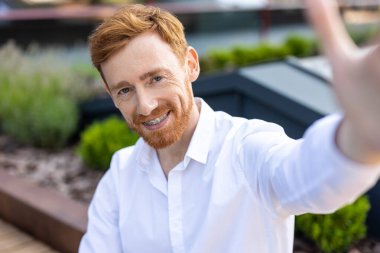 Beyaz tişörtlü kızıl sakallı adam pov selfie çekiyor. Kentin sokaklarında kendine güveni tam duruyor.