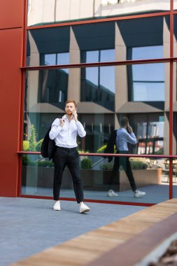 Beyaz tişörtlü kızıl saçlı adam omzunun üzerinde ceketini tutuyor ve modern şehir ortamında akıllı telefondan konuşuyor.