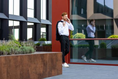 Şık kızıl saçlı, resmi giyinmiş, iş alanında yürürken akıllı telefondan iletişim kuruyor.