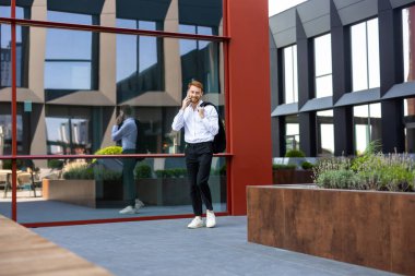 Sakallı, beyaz gömlekli, ceketli, ofis binasının önünde konuşan kızıl saçlı adam.