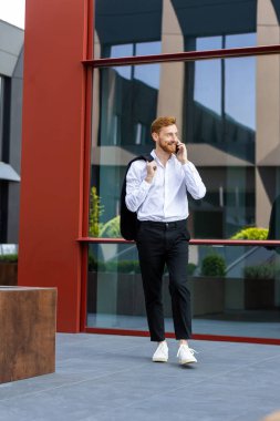 Modern şirket bölgesinde yürürken cep telefonuyla konuşan kızıl saçlı profesyonel bir adam.