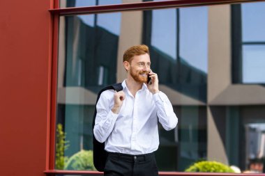 Kızıl saçlı, şık giyimli, ceketli ve şehir merkezinde telefonla konuşan bir adam.
