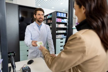 Eczanede. Genç, siyah saçlı, beyaz kadın eczaneden ilaç alıyor.