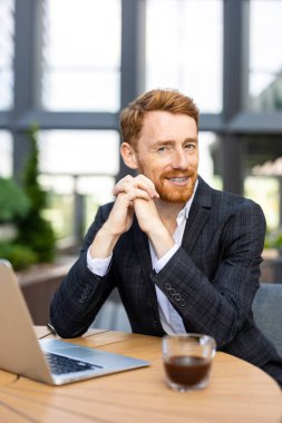 Takım elbiseli kendine güvenen genç adam, kafeterya masasında dizüstü bilgisayarla poz veriyor.