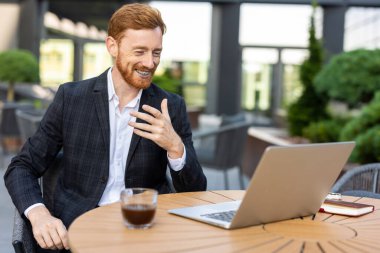 Takım elbiseli genç zencefilli işadamı açık kafede görüntülü görüşme yapıyor.