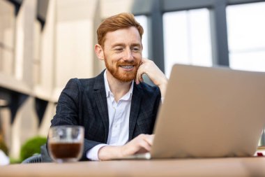 Neşeli kızıl sakallı profesyonel adam dışarıda bilgisayar başında poz veriyor.