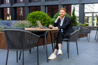 Neşeli kızıl sakallı profesyonel adam dışarıda bilgisayar başında poz veriyor.