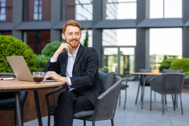 Modern ticaret bölgesindeki açık masa masasında dizüstü bilgisayar kullanan sakallı kızıl saçlı.