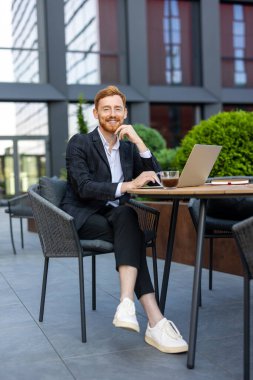 Gülümseyen kızıl saçlı iş adamı güneşli bir günde laptopla açık kafede çalışıyor.