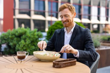 Kızıl saçlı işadamı, şehirdeki açık kafe masasında öğle yemeğinin tadını çıkarıyor.