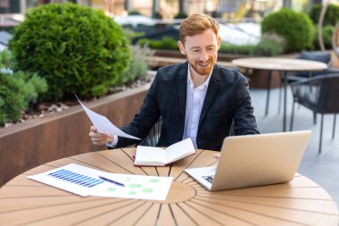 Kızıl saçlı işadamı dışarıda belgelerle birlikte dizüstü bilgisayarda çalışıyor.