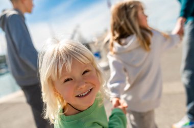 Sarışın çocuk marinada dikilirken kameraya gülümsüyor arkasında bulanık insanlar ve yatlar var.