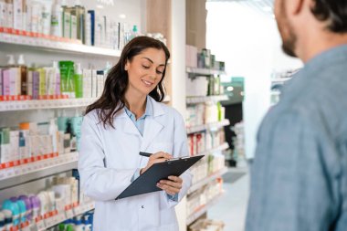 Esmer kadın eczacı, eczanedeki müşteriye danışmanlık yaparken not alıyor.