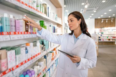 Eczane raflarındaki ürünlere pano ile bakan esmer kadın eczacı.